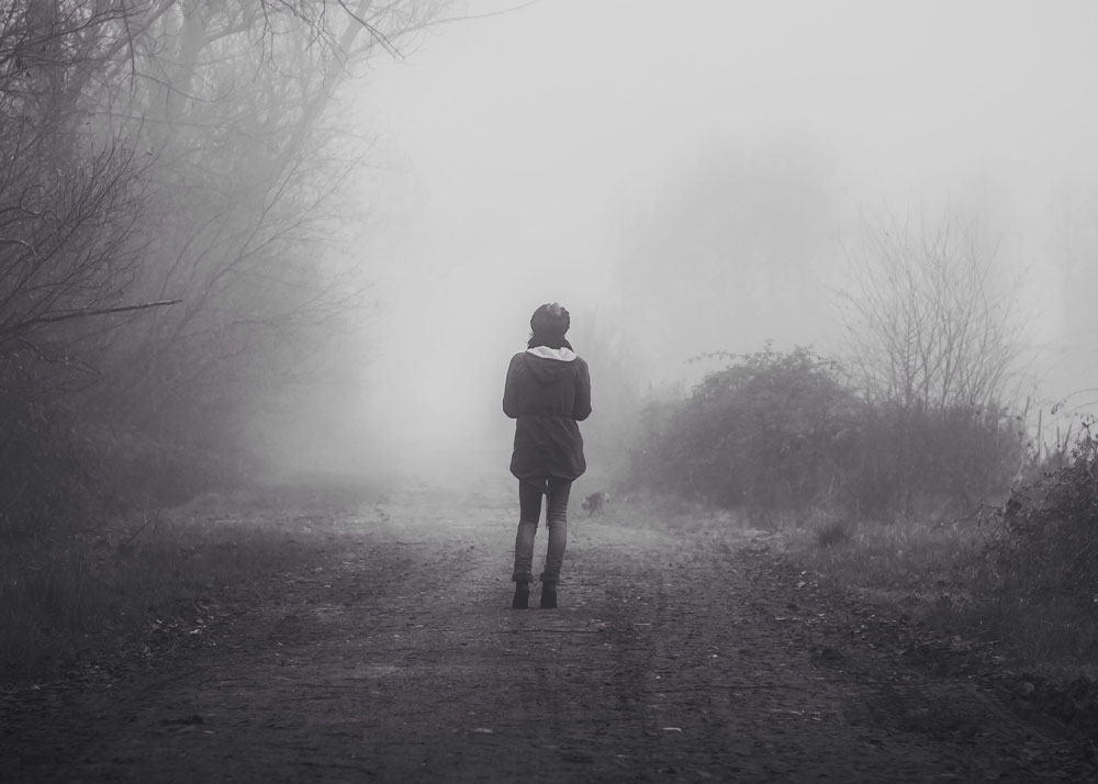 A girl walks down a dirt road in the fot. It is autumn or winter, and the trees are bare of leaves. She wears a jacket.