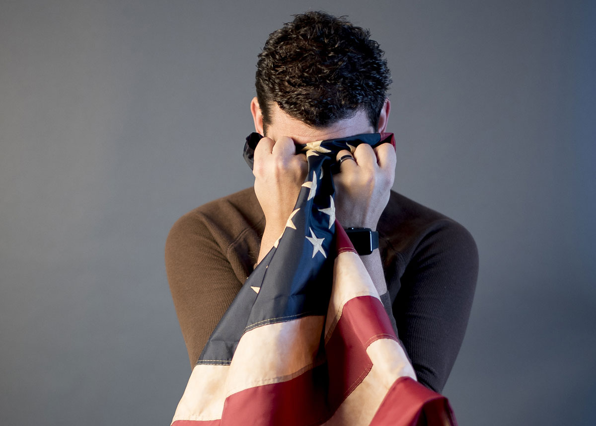 A man with curly dark hair holds an American flag over his face in a state of grief